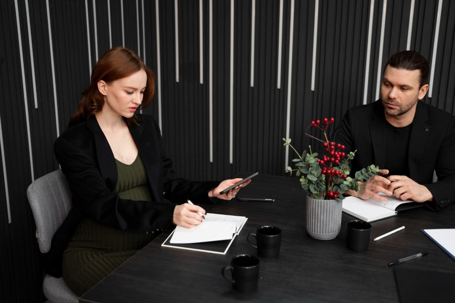 A woman writes notes while checking her phone at a black conference table adorned with a vase of red berries, as a man listens attentively. Captures the importance of cybersecurity for law firms and managed IT services in maintaining compliance during internal meetings.