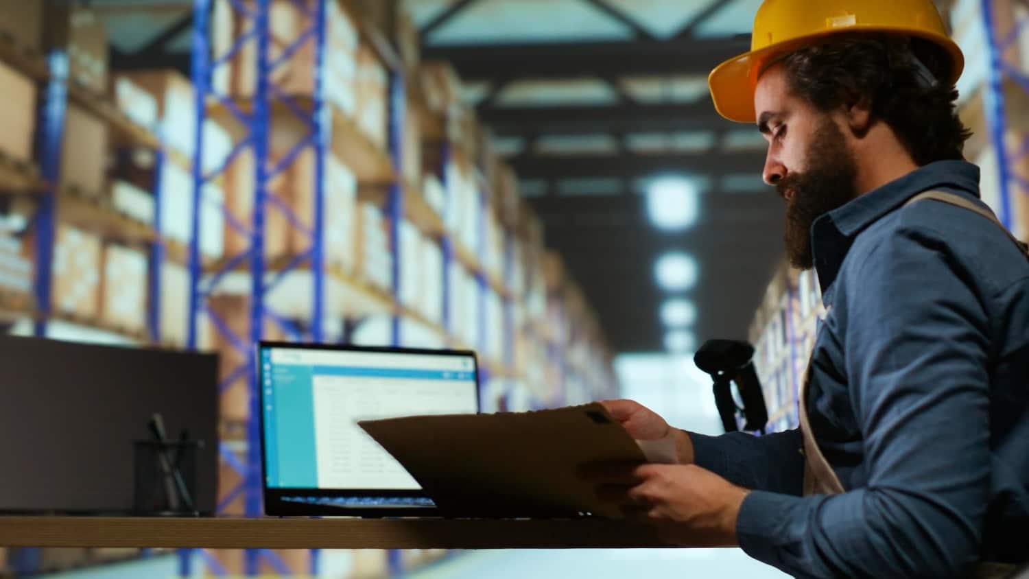 A male worker in a yellow hard hat sits at a desk in a warehouse, reviewing files next to an open laptop displaying software. This image conveys industrial IT infrastructure and cybersecurity for manufacturing in logistics or inventory tracking systems.