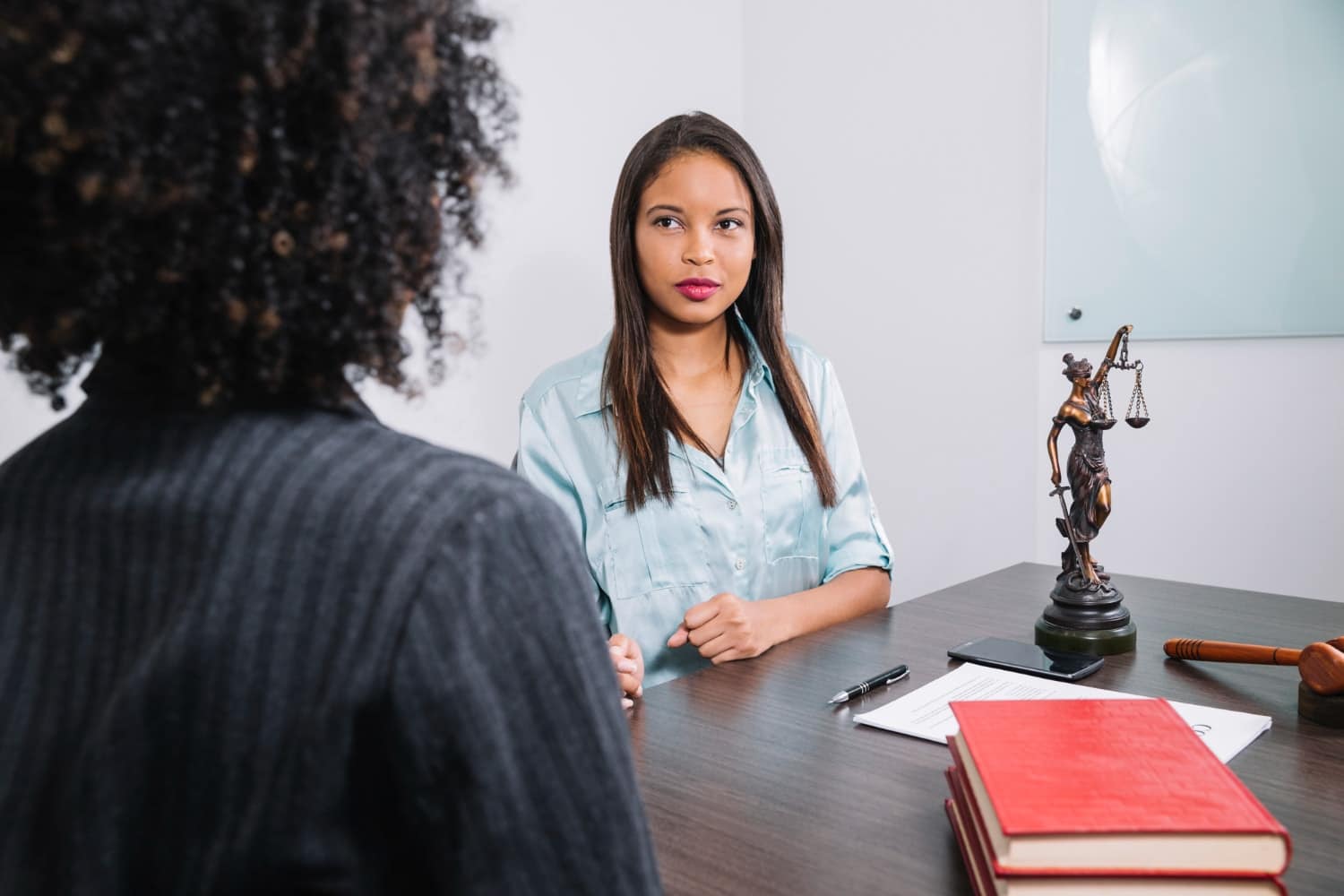 Two women sit across a desk discussing legal matters; a Lady Justice statue, legal documents, and law books are visible on the table. Highlights cybersecurity for law firms and managed IT services that safeguard confidential legal data.