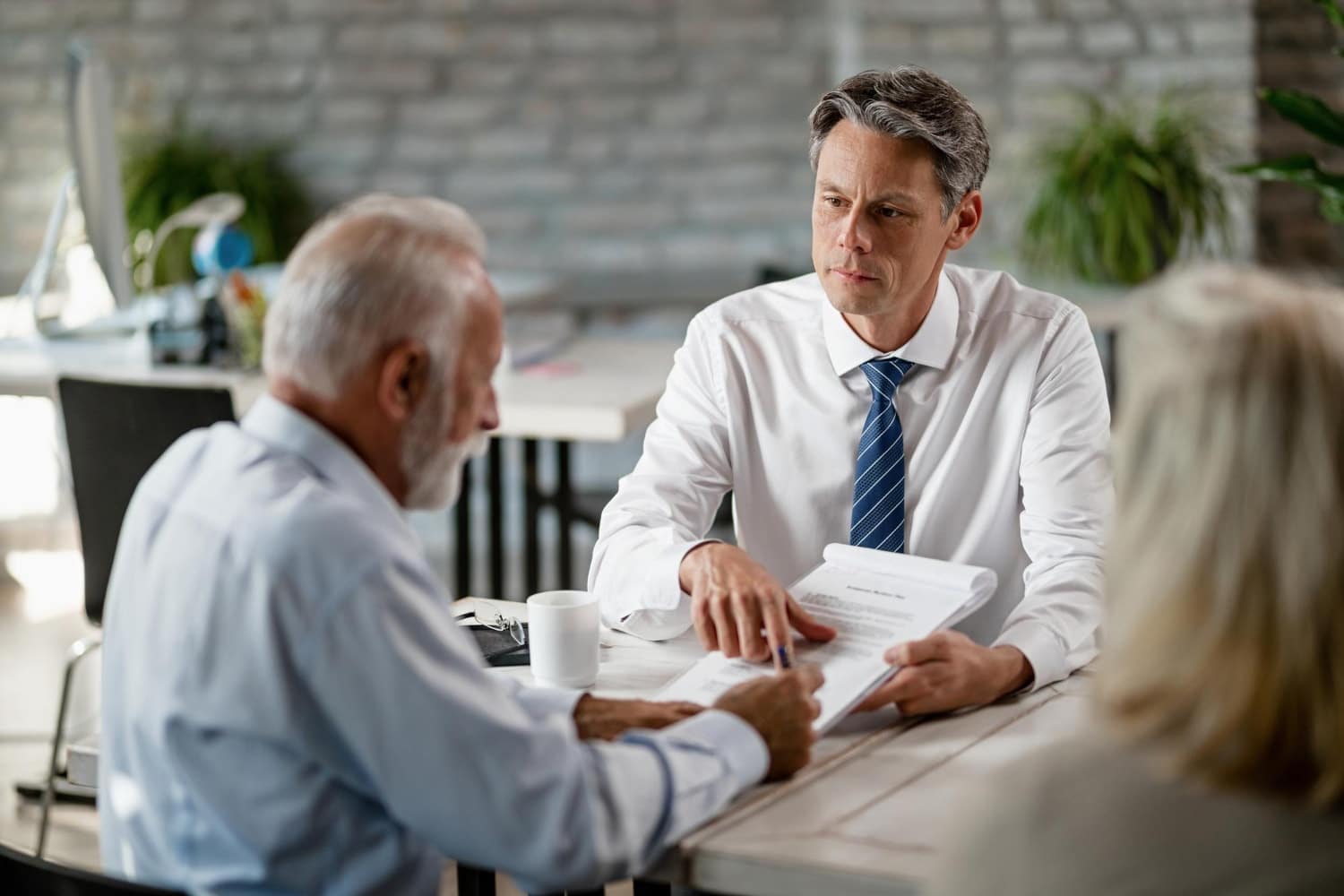 A serious conversation between two men and a woman at a table, with one man explaining a document to an elderly client. Highlights cybersecurity for law firms and managed IT services that ensure secure handling of legal agreements.
