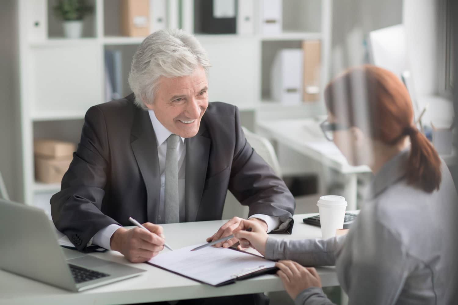 An older man in a suit smiles while signing documents during a discussion with a woman in a bright, modern office. Depicts IT services for law firms, focusing on user-friendly IT support services and managed IT services for client satisfaction.