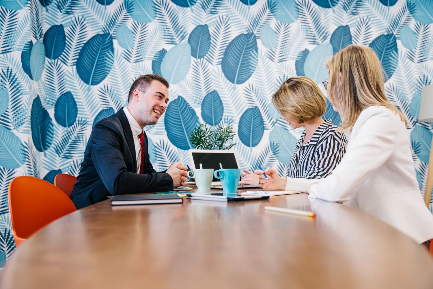 A man in a suit smiles while meeting with two women in a bright room with a blue leaf-patterned wall, with laptops and coffee mugs on the table. Illustrates IT services for law firms, managed IT services, and client-focused technology support.