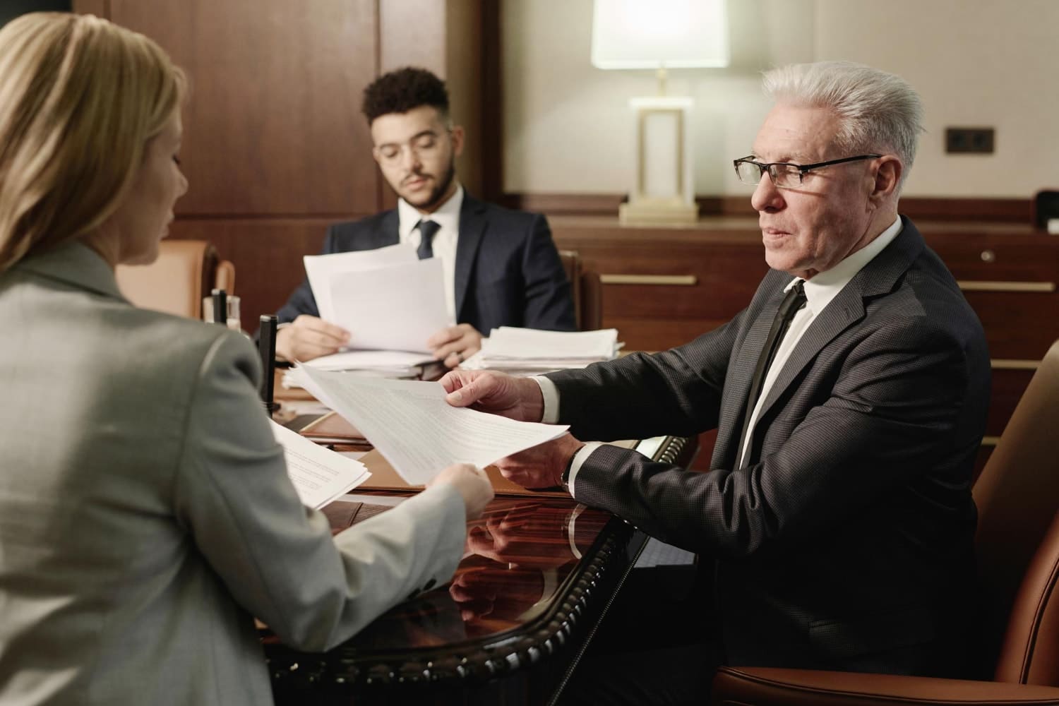 An older man in glasses reads legal documents across from a woman at a polished conference table, while another man reviews papers in the background. Represents legal IT compliance, IT support services, and secure documentation practices for law firms.