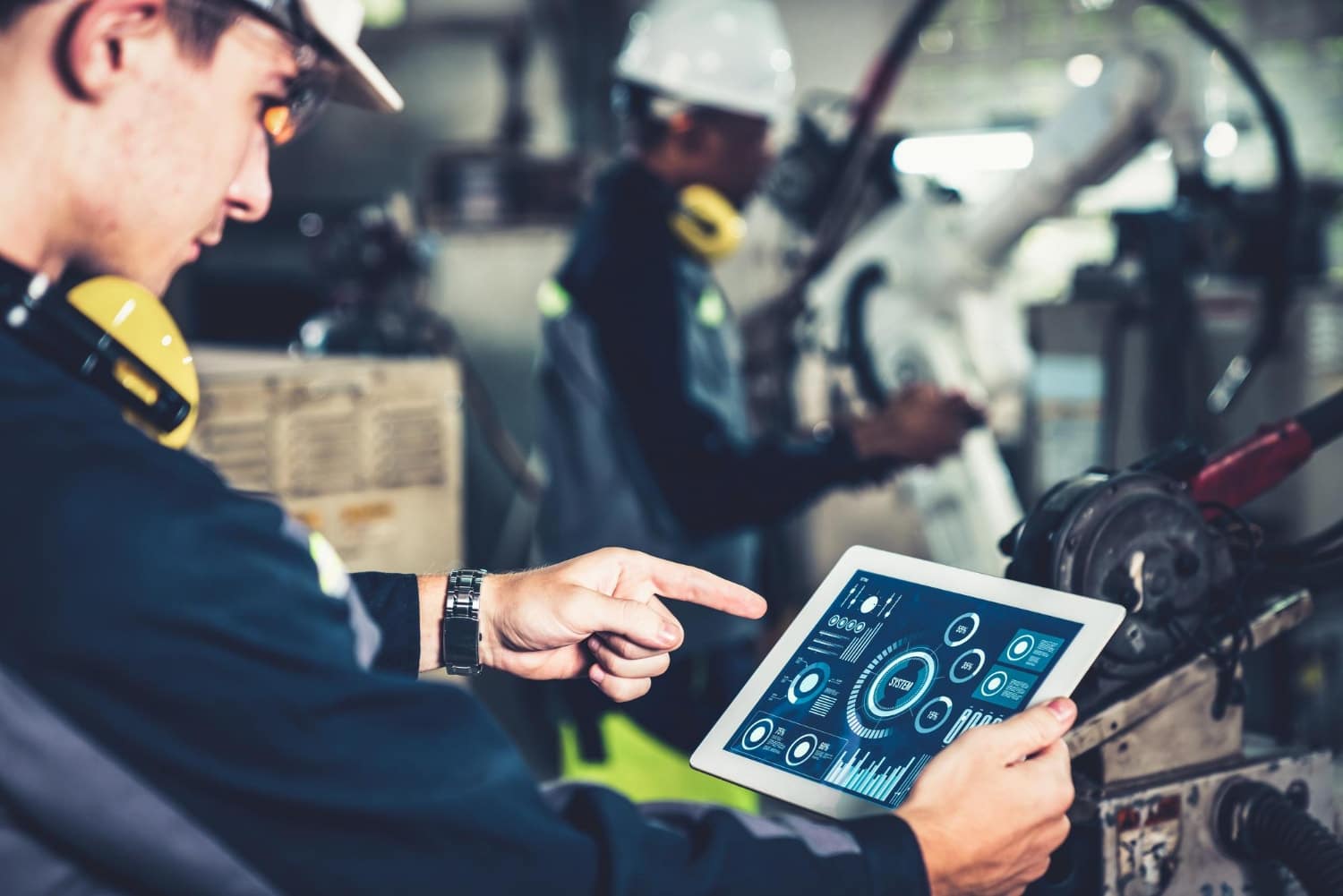 An industrial technician uses a tablet to control or monitor machinery with digital dashboards displayed on-screen. This image demonstrates IT solutions for manufacturing and cybersecurity for manufacturing through real-time system diagnostics.