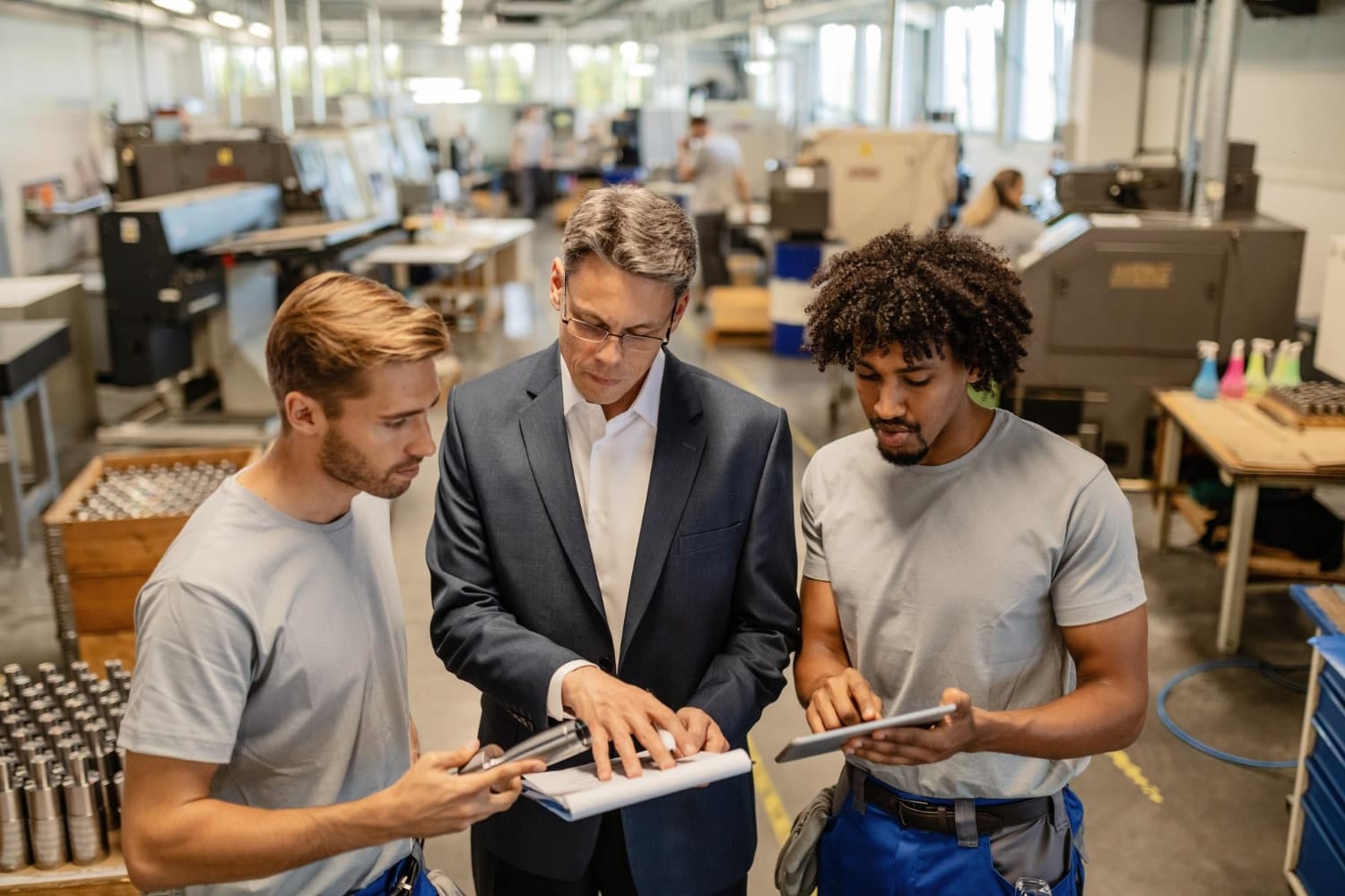 A man in a business suit discusses documents with two factory employees holding digital tablets and tools. The setting illustrates managed IT services and IT solutions for manufacturing through digital collaboration and operational support on the factory floor.
