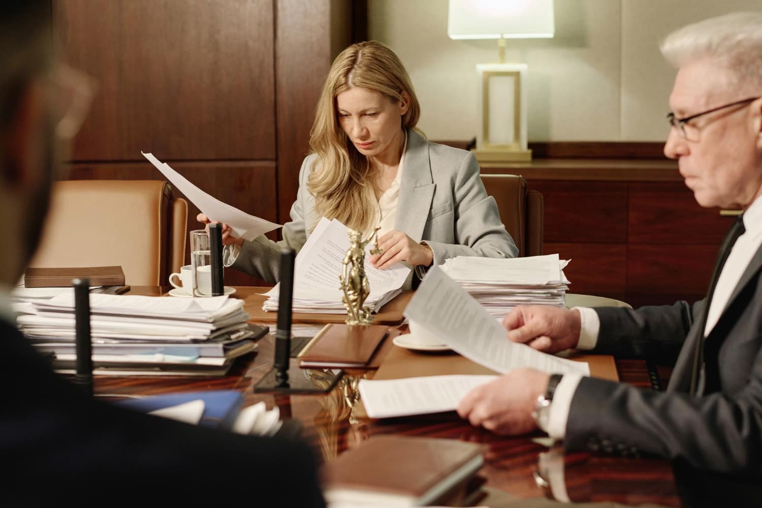 A woman in a gray blazer reviews legal documents at a large conference table piled with papers, alongside two men in suits. Symbolizes IT support services and legal IT compliance for law firms handling sensitive client data.