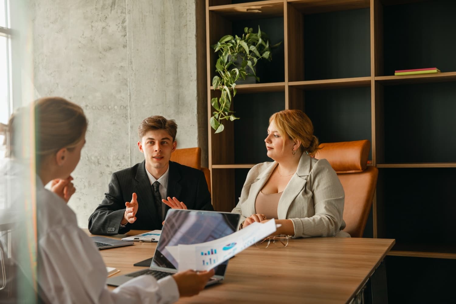 A young man in a suit gestures while speaking to two women in a meeting room with laptops and charts. Depicts IT support services and managed IT services for law firms, emphasizing collaborative technology solutions.